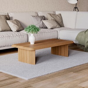 K&ouml;nnte beinhalten: Ein heller Holz-Couchtisch mit abgerundeter Kante und einer wei&szlig;en Vase mit einer kleinen gr&uuml;nen Pflanze darauf. Der Tisch steht auf einem wei&szlig;en Teppich vor einem wei&szlig;en Sofa.