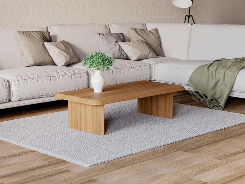 K&ouml;nnte beinhalten: Ein heller Holz-Couchtisch mit abgerundeter Kante und einer wei&szlig;en Vase mit einer kleinen gr&uuml;nen Pflanze darauf. Der Tisch steht auf einem wei&szlig;en Teppich vor einem wei&szlig;en Sofa.