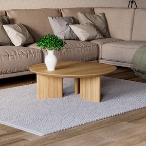 May include: A modern living room scene featuring a light brown sectional sofa with several pillows, a light wood oval coffee table, a gray rug, and a white vase with greenery. The room has a neutral color palette.