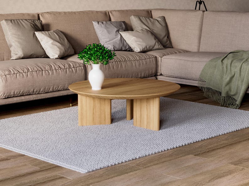 May include: A modern living room scene featuring a light brown sectional sofa with several pillows, a light wood oval coffee table, a gray rug, and a white vase with greenery. The room has a neutral color palette.