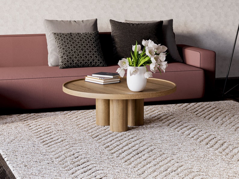 May include: A light brown wooden coffee table with a round top and four cylindrical legs. The table is decorated with a white vase filled with white tulips and a stack of books. The table is sitting on a cream-coloured rug with a wavy pattern.