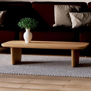 Könnte beinhalten: Ein heller Holz-Couchtisch mit abgerundeter Kante und zwei Beinen auf jeder Seite. Der Tisch steht auf einem hellgrauen Teppich vor einem braunen Ledersofa mit Kissen.