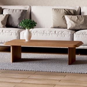 K&ouml;nnte beinhalten: Ein heller Holz-Couchtisch mit einer rechteckigen Tischplatte und zwei quadratischen Beinen auf jeder Seite. Der Tisch steht auf einem hellgrauen Teppich vor einem wei&szlig;en Sofa mit Kissen.