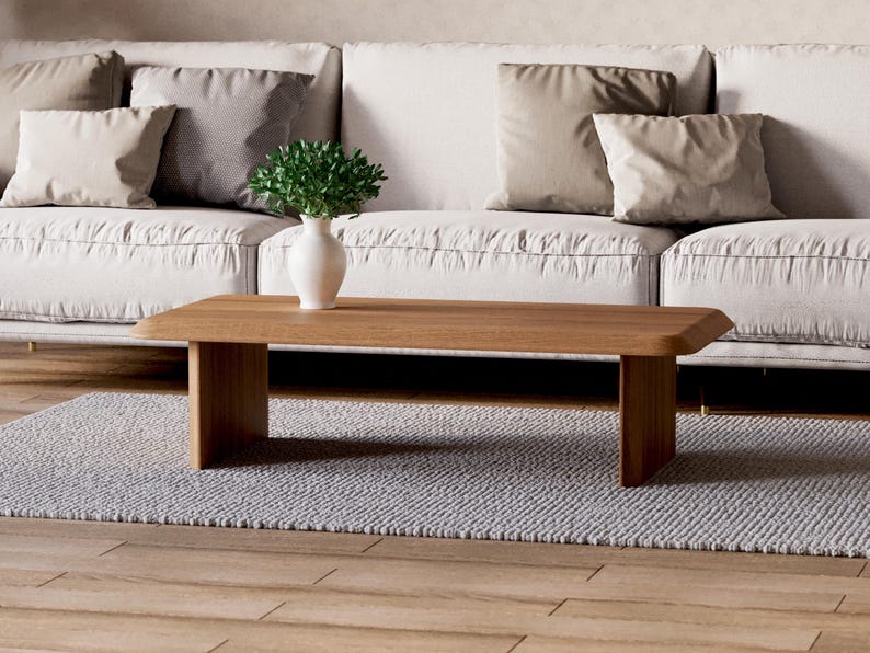 K&ouml;nnte beinhalten: Ein heller Holz-Couchtisch mit einer rechteckigen Tischplatte und zwei quadratischen Beinen auf jeder Seite. Der Tisch steht auf einem hellgrauen Teppich vor einem wei&szlig;en Sofa mit Kissen.