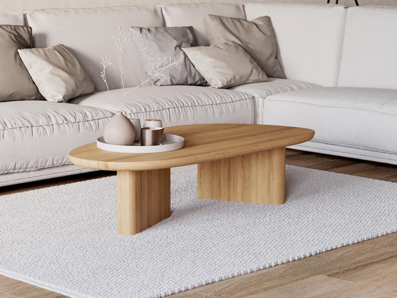 Puede incluir: Una mesa de centro ovalada de madera clara con dos patas rectangulares, sobre una alfombra blanca texturizada. Un sof&aacute; blanco con cojines beige est&aacute; en el fondo. Una bandeja blanca con objetos decorativos est&aacute; sobre la mesa.
