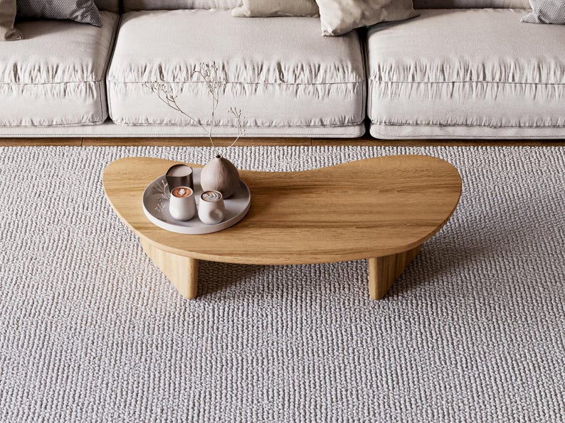 May include: A wooden coffee table with a distinctive, curved shape. The table has three legs and a tray with decorative items, including small cups and a vase. The table sits on a light grey rug, with a white sofa in the background.