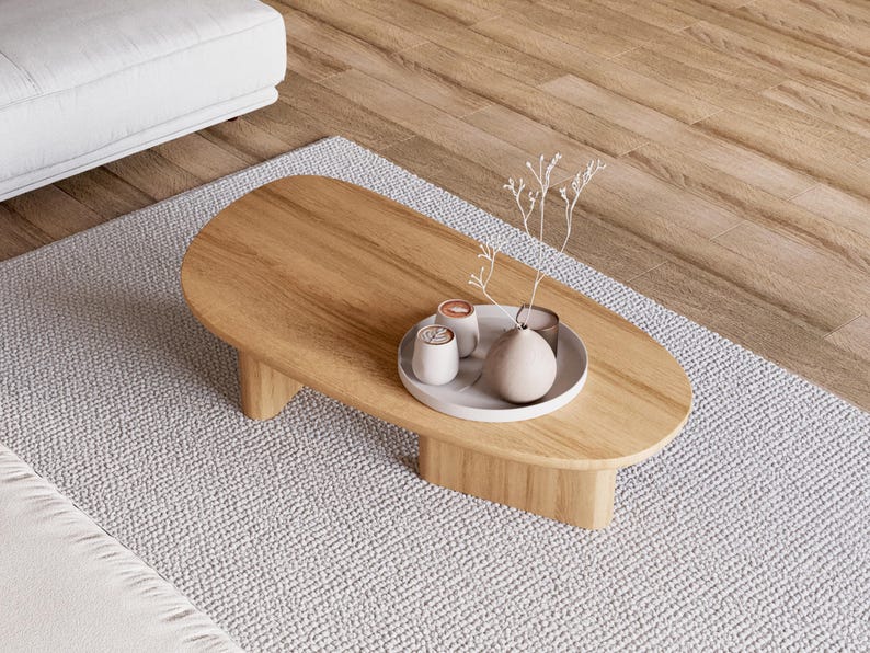 Puede incluir: Una mesa de centro ovalada de madera marr&oacute;n claro, con una base clara. Una bandeja blanca en la parte superior contiene objetos decorativos, incluyendo un jarr&oacute;n y peque&ntilde;as tazas. La mesa descansa sobre una alfombra gris. La mesa mide unos 120 cm de largo.