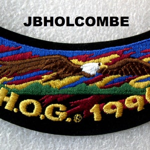 Può includere: Una toppa da motocicletta ricamata in nero e oro con un'aquila in volo su uno sfondo rosso, giallo, blu e verde. La toppa riporta la scritta "H.O.G. 1996" e "JBHOLCOMBE".