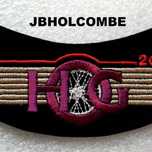 Può includere: Una toppa da moto nera con un design ricamato rosso e bianco. La toppa presenta le lettere "H.G." con una ruota al centro e il testo "2001" sopra il design. Il testo "JBHOLCOMBE" è stampato sopra la toppa.