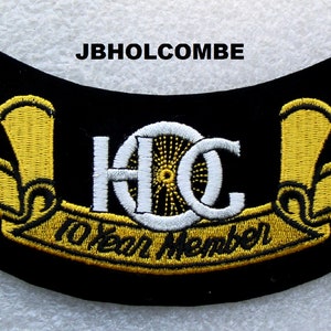Puede incluir: Un parche bordado negro con letras doradas y un diseño de rueda blanca. El parche dice "H.C. 10 Year Member".