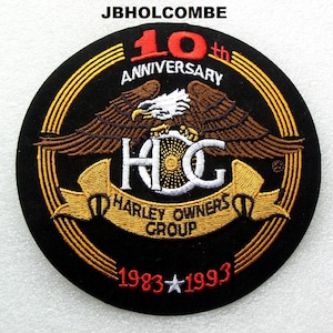 May include: A black, circular patch embroidered with the text "10th Anniversary" in red and gold. It features a detailed eagle with outstretched wings above the letters "HOG" and a banner reading "Harley Owners Group". The years "1983-1993" are also displayed.