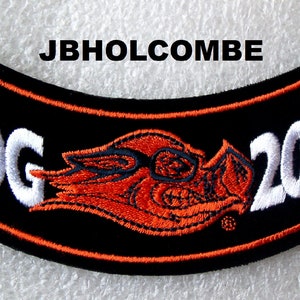Könnte beinhalten: Schwarzer und orangefarbener Motorrad-Patch mit weiß gesticktem Text "HOG" und "2006" sowie einem rot und schwarz gestickten Schweine-Logo.