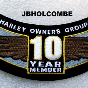 May include: Black, embroidered patch with the text "HARLEY OWNERS GROUP" and "10 YEAR MEMBER". The patch features a wing design in brown and a light blue background. The text "JBHOLCOMBE" is at the top.