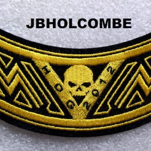 May include: A black and gold motorcycle patch with a skull and crossbones design. The patch has the text "MC" and "2012" on either side of the skull. The text "JBHOLCOMBE" is above the patch.