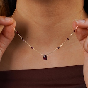 May include: A delicate gold necklace featuring small, round, dark red gemstones and a teardrop-shaped pendant. The necklace is being held up, showcasing its design and the way it lays on the neckline. The necklace is a fine piece of jewelry.