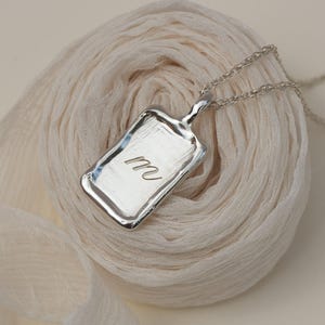 Op de afbeelding: Een zilverkleurige rechthoekige hangerketting met de letter "M" gegraveerd. De hanger hangt aan een delicate ketting en rust op een crème-kleurige stof. De hanger heeft een getextureerde, onregelmatige rand.
