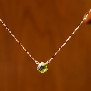 May include: A delicate gold necklace featuring a pear-shaped, light green peridot gemstone pendant. The pendant is suspended from a fine gold chain. The background is a warm, brown tone, highlighting the jewelry.