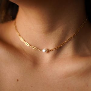 May include: A gold-colored chain necklace with a single white pearl pendant. The necklace has a link chain design and is worn around the neck. The background is blurred, focusing attention on the jewelry.