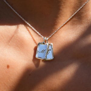Op de afbeelding: Een zilveren ketting met twee rechthoekige hangers. Elke hanger heeft een witte achtergrond met een cursieve initiaal in zwart. De hangers zijn verbonden en hangen aan een fijne zilveren ketting.