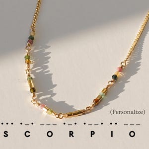 以下が含まれることがあります： カラフルな宝石ビーズが付いたゴールドトーンのネックレス。繊細なチェーンと円筒形のゴールドアクセントが特徴です。「SCORPIO」の文字がネックレスの下にモールス信号で表示され、括弧内には「パーソナライズ」という文字があります。