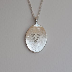 Puede incluir: Collar con colgante plateado. El colgante ovalado tiene una superficie texturizada y la letra "V" grabada en el centro. La cadena es de diseño delicado y retorcido. Una joya sencilla y elegante.