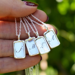 Op de afbeelding: Zilveren kettingen met rechthoekige hangers, elk met een initiaal in sierlijke letters: J, M, L en A. De hangers hangen aan fijne zilveren kettingen, tegen een vage groene achtergrond.
