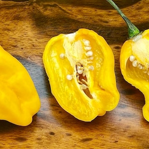 May include: Three yellow habanero peppers, one whole and two halved, showing the seeds inside. The peppers are on a wooden surface.