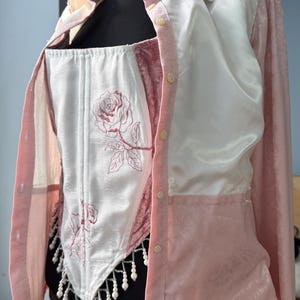 May include: A two-piece outfit comprising a white corset with rose embroidery and a pink button-down shirt. The shirt has a satin finish and a collared neckline. The corset has a beaded fringe.