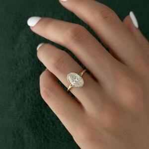 May include: A gold ring with an oval-shaped diamond. The diamond is set in a bezel setting, and the band is a thin, polished gold. The ring is on a finger, with a dark green background.
