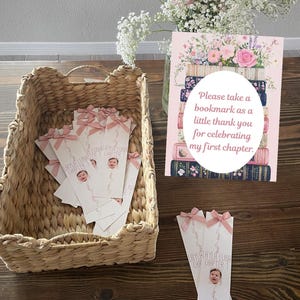 May include: A woven basket filled with white bookmarks, each adorned with a pink bow and a baby's photo. A sign with floral illustrations and text that reads "Please take a bookmark as a little thank you for celebrating my first chapter."