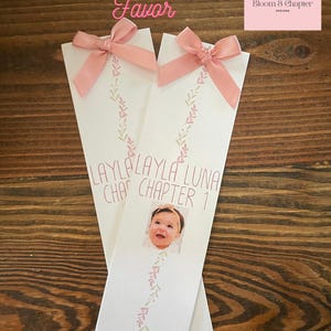 May include: Two white bookmarks with pink bows at the top. Each bookmark has a photo of a baby, the name "LAYLA LUNA", and the text "CHAPTER 1". The bookmarks are decorated with pink and green floral designs. The words "Editable Bookmark Favor" are in pink script.