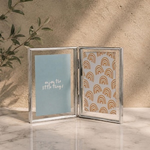 May include: A silver-toned hinged double picture frame with clear glass panels. The left panel contains a light blue insert with the white text "enjoy the little things," while the right panel features a pattern of repeating yellow rainbows.