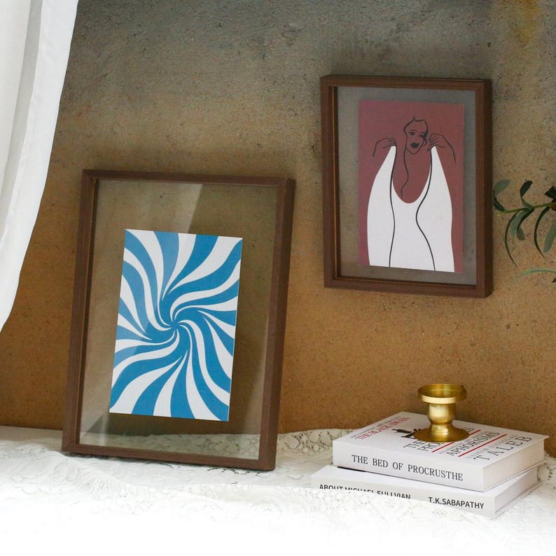 May include: Two rectangular brown wooden frames with glass panels. One frame contains a blue and white spiral pattern print. The other frame displays a line art illustration of a figure in a white garment against a muted red background.