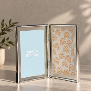 May include: A double-hinged silver metal picture frame with two glass panels. The left panel features a light blue background with the white text "enjoy the little things," while the right panel displays a repeating pattern of orange rainbows.