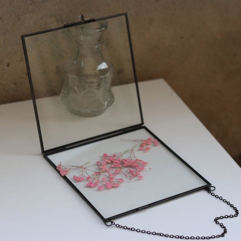 May include: A black metal frame with a glass front and back, holding dried pink flowers. The frame has a chain attached to the top for hanging.