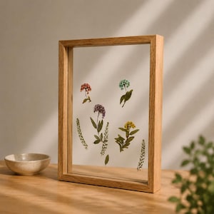 May include: A rectangular light-toned wooden floating frame containing several pressed dried flowers in shades of red, purple, green, and yellow. The frame features two clear glass panes that hold the botanical specimens in a vertical arrangement.