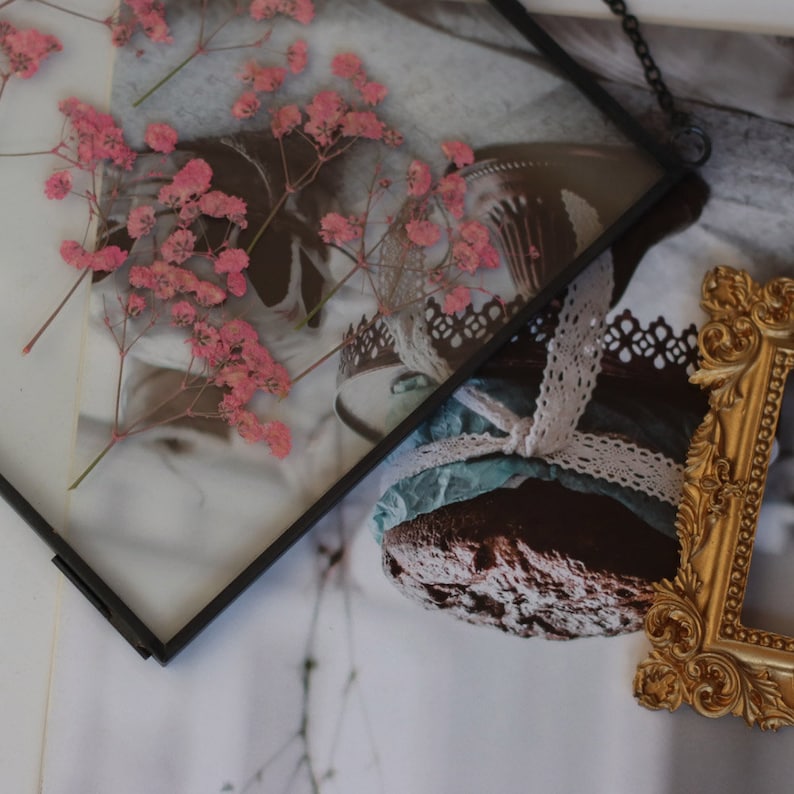 May include: A black framed picture with dried pink flowers and a vintage-style cake with lace and blue ribbon. The picture is sitting on a white surface next to a gold ornate picture frame.