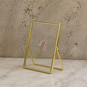 Può includere: Una cornice portafoto rettangolare in metallo dorato con un bordo sottile e perlato. La cornice presenta due lastre di vetro trasparente tenute insieme dal bordo metallico, con un fiore pressato rosa e bianco visibile tra i vetri.