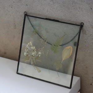 May include: A square black metal frame with a glass front, holding pressed flowers and leaves. The frame has a chain hanging from the top.