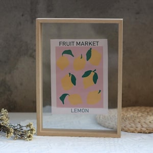 Op de afbeelding: Een houten fotolijst met een heldere glazen voorkant, waarop een roze en gele print van citroenen is afgebeeld met de tekst "FRUIT MARKET" en "LEMON".