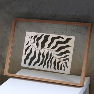 Puede incluir: Una impresión enmarcada en blanco y negro de una hoja estilizada con un tallo curvo y líneas onduladas. La impresión está sobre un fondo crema y está enmarcada en un marco de madera natural.