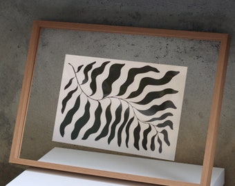 May include: A rectangular light wood frame containing a clear glass pane. Centered within the frame is a white rectangular print featuring a black graphic illustration of a single long, wavy, fern-like leaf with multiple curved segments.