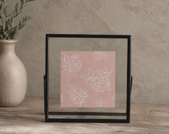 Puede incluir: Un marco flotante cuadrado de metal negro que contiene una lámina de color rosa empolvado con un patrón floral de líneas blancas. El marco presenta un borde fino y está diseñado para exhibir obras o fotos entre dos paneles de vidrio.