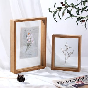 以下が含まれることがあります： ガラス製の前面を持つ木製のフォトフレームが2つ。大きなフレームは左側にあり、押し花が入っています。小さなフレームは右側にあり、乾燥した花の枝が入っています。両方のフレームは白い表面の上に置かれています。
