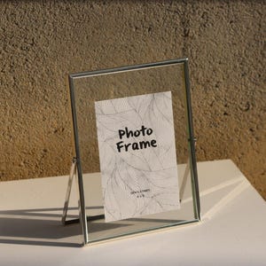 May include: A silver photo frame with a clear glass front and a stand. The frame is sitting on a white surface. The text "Photo Frame" is written on a white card inside the frame.
