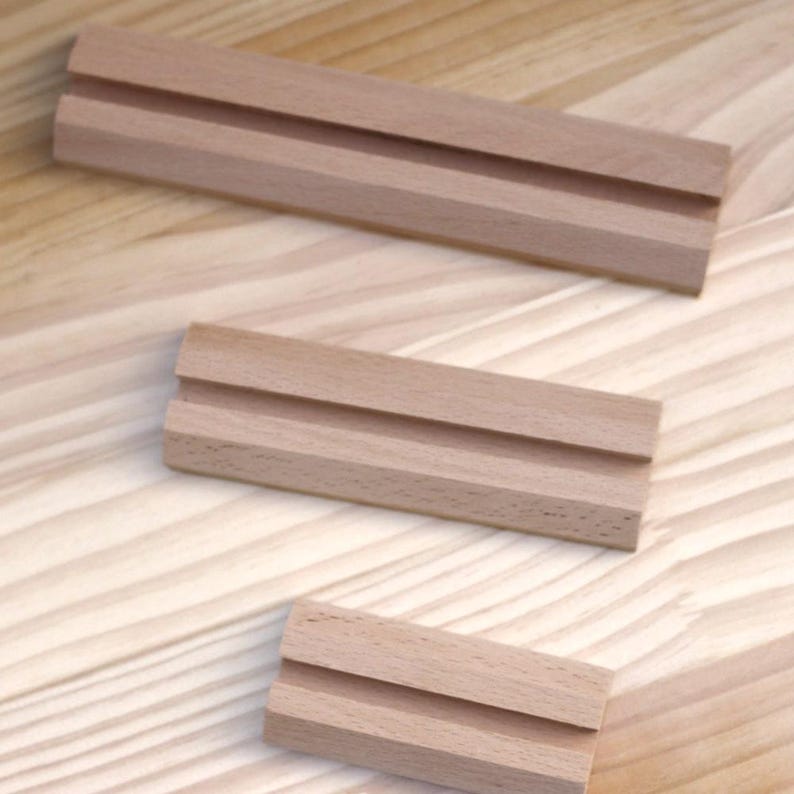 May include: Three wooden display stands with a groove in the center. The stands are made of light brown wood and are arranged in a staggered pattern.