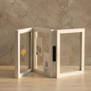 May include: A white, three-panel folding picture frame with clear glass panes. The left panel holds a yellow ginkgo leaf, the center panel contains two white cards with black floral illustrations, and the right panel displays a gold fern leaf.