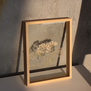 Op de afbeelding: Een houten frame met een heldere glazen inzet, waarin een geperste bloemenarrangement van witte en lichtroze bloemen met groene stelen te zien is. Het frame staat op een lichtbruin oppervlak.