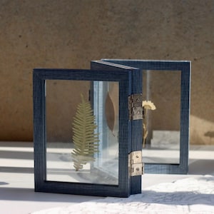 May include: A folding double picture frame with a dark blue wood grain finish and clear glass panels. The frame is hinged with silver metal hardware and contains a pressed green fern leaf, a brown leaf, and a small yellow flower inside.