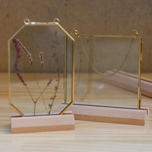May include: Two gold-toned glass picture frames with wooden bases. The frame on the left is octagonal and contains dried flowers. The frame on the right is square and has a chain hanging from the top.
